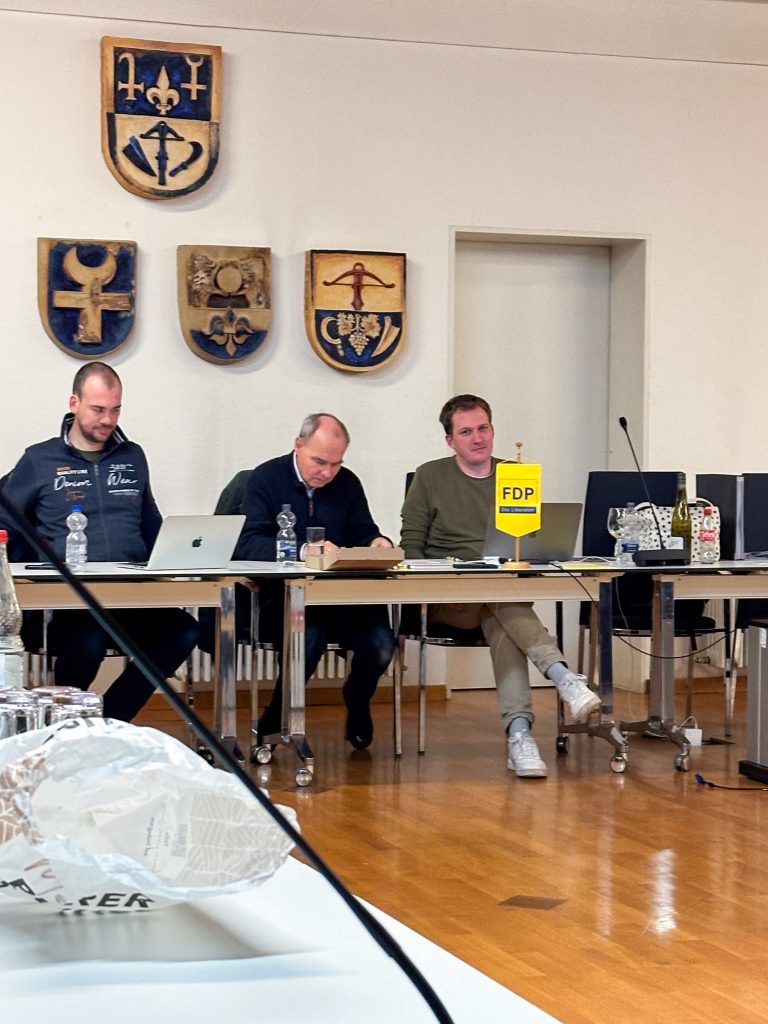 Menschen bei Versammlung mit Wappenschildern im Hintergrund.